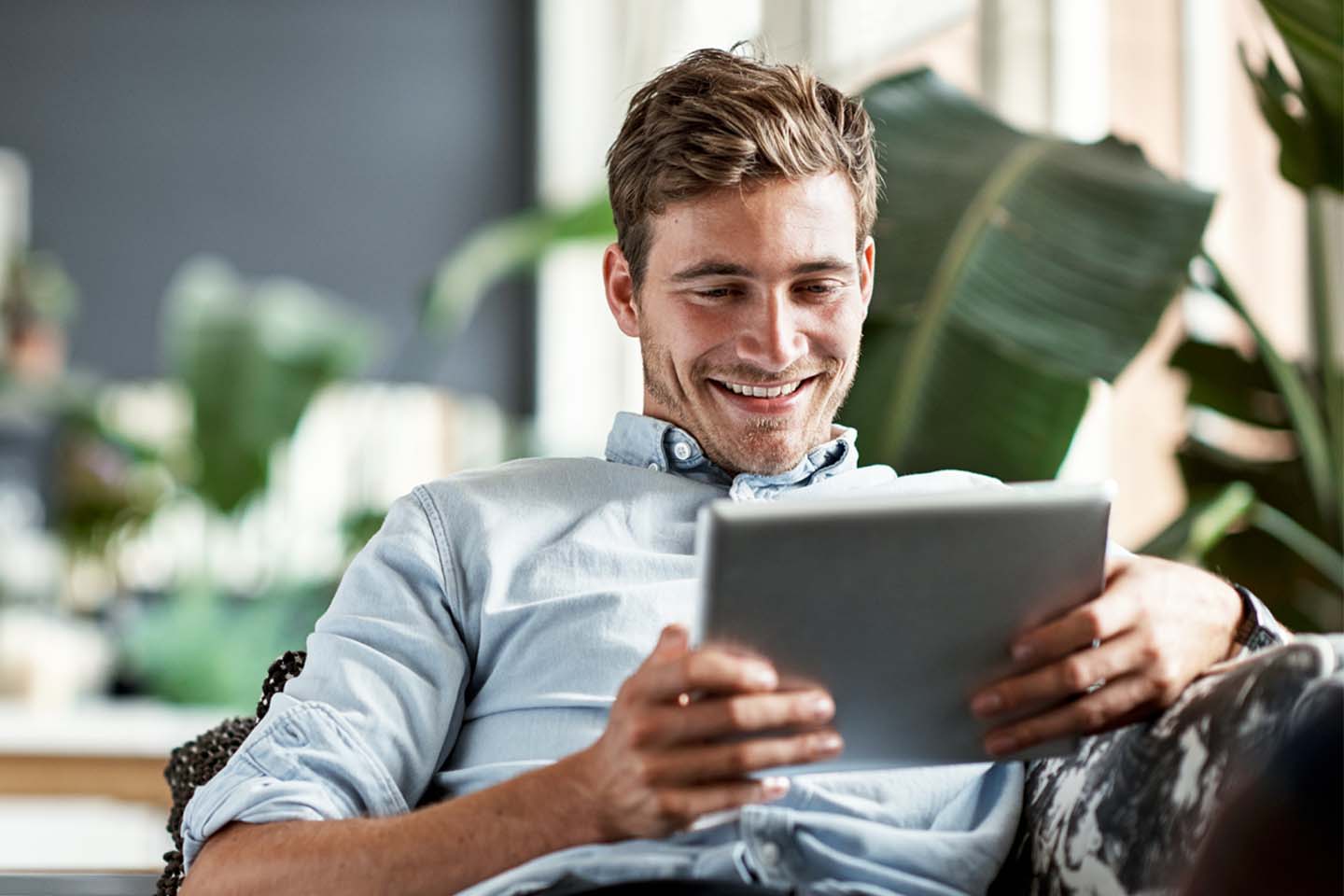 Ein junger Mann mit Dreitagebart sitzt lässig auf einem Sofa. Er ist umgeben von hohen Grünpflanzen und schaut lächelnd auf seinen Tablet-Computer. 