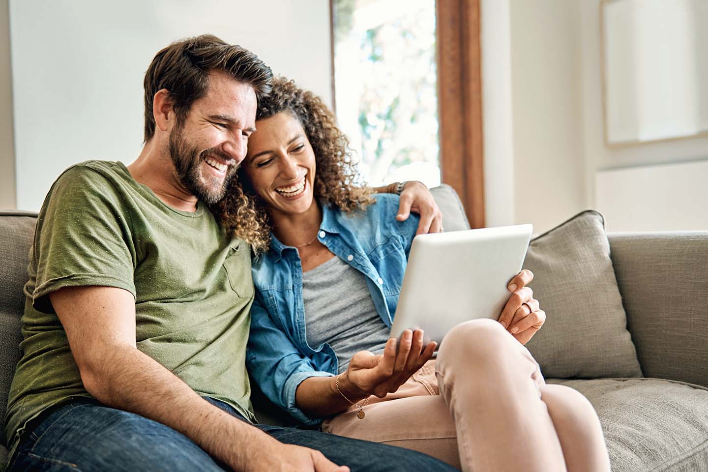 Ein junges Paar sitzt eng aneinander gelehnt lächelnd auf dem Sofa. Er trägt einen Vollbart, sie hat stark gelockte, braune Haare. Beide sind freizeitlich angezogen und blicken gemeinsam auf einen Tablet-Computer.